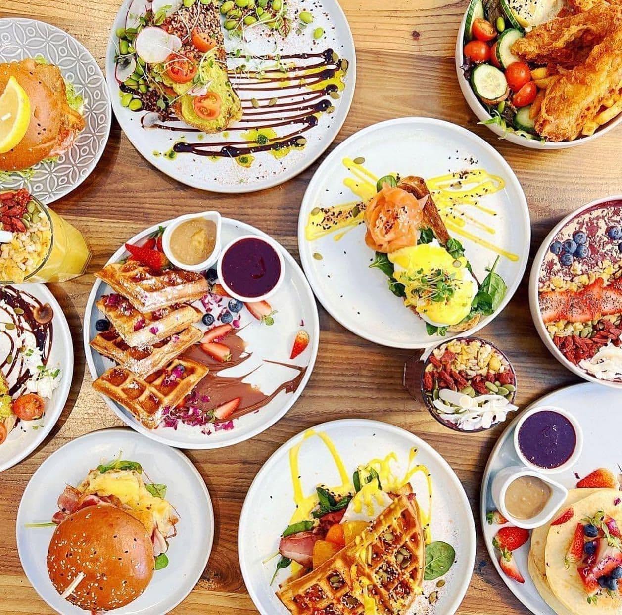 A table of breakfast plates seen from above — waffles, eggs benedict, burgers and smoothie bowls