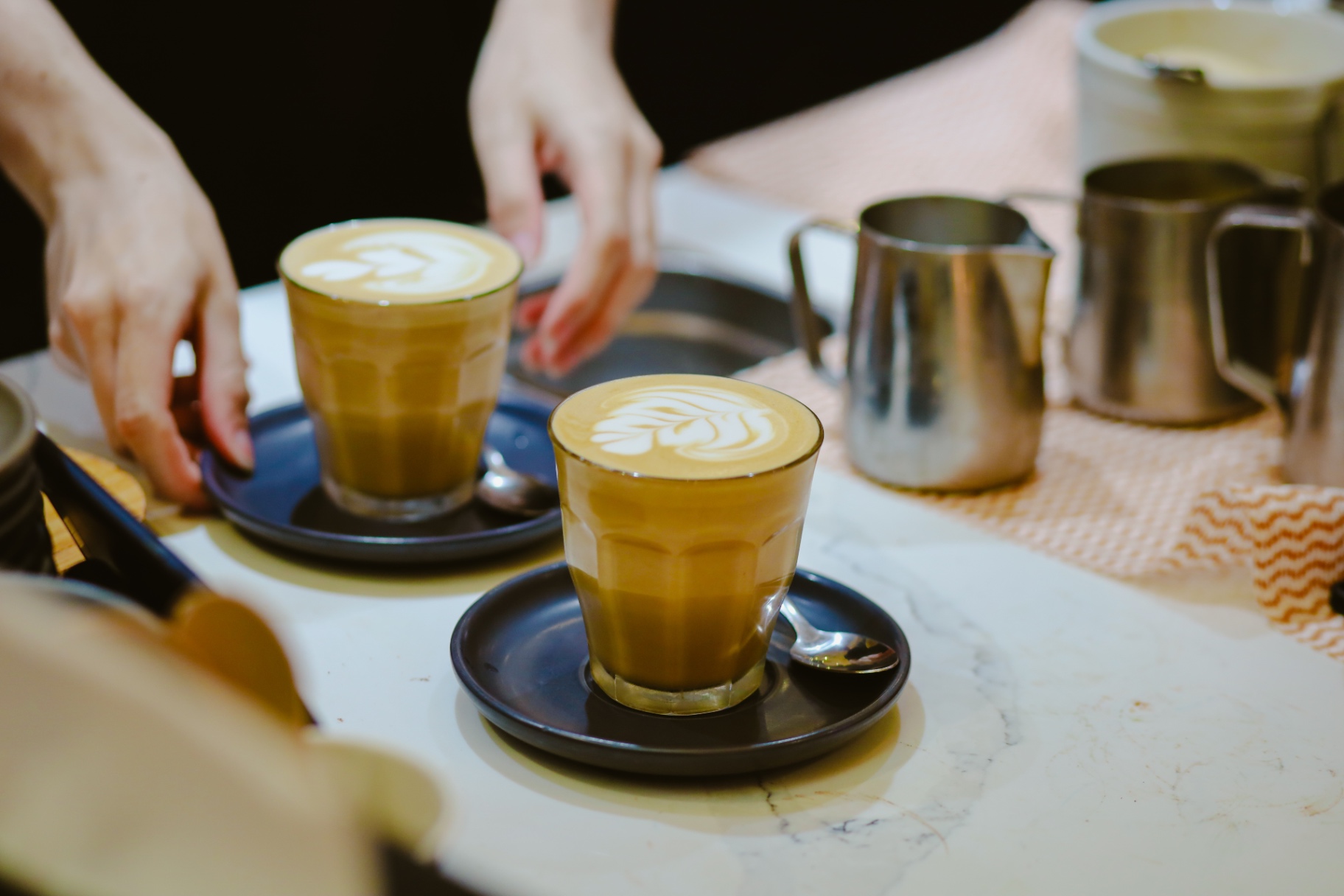Two lattes with latte art being set down on a marble counter beside steel milk jugs