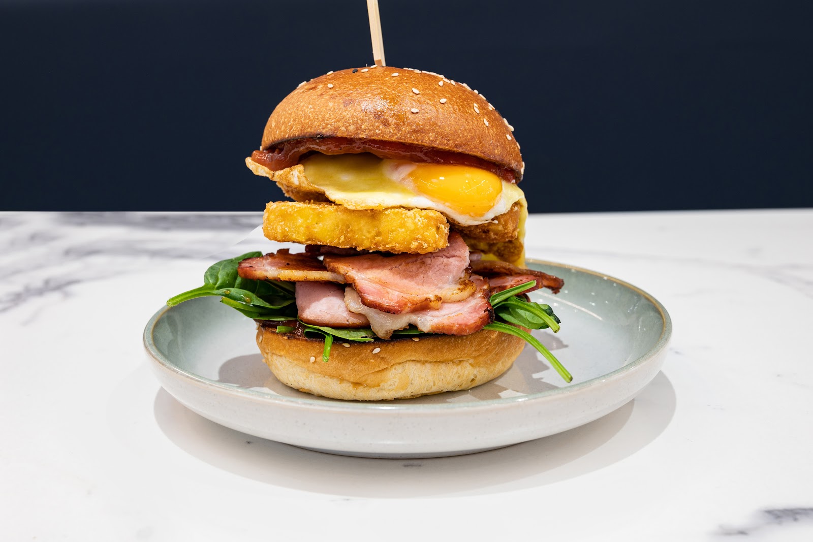 A loaded breakfast burger — bacon, fried egg with a runny yolk, hash brown and spinach on a sesame bun
